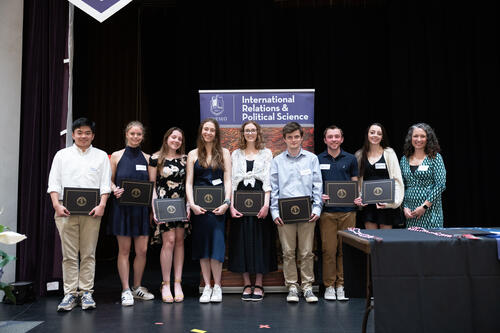 Eight students and a professor stand on a stage holding their awards certificates.